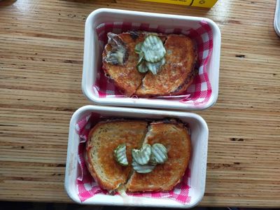A photo of two cheese toasties with pickles on top in takeaway containers