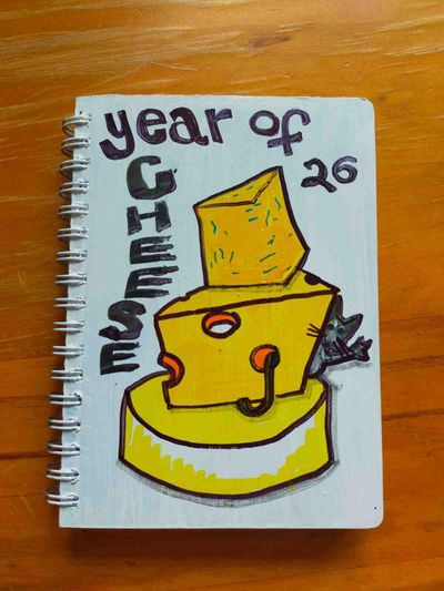 A diary cover painted white with a picture of a stack of various types of cheese, with a mouse waving from behind it. The cover says 'year of CHEESE 26'.